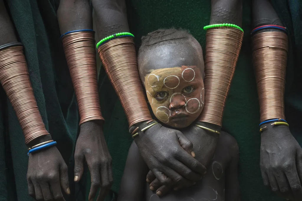 Nejlepší single fotografie z portfolií v kategorii Tváře, lidé a kultura. Kibish, Etiopie. Chlapec obklopen měděnými náramky žen, tradičními doplňky kmene Suri