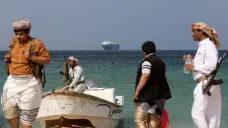 Ozbrojení muži stojí na pobřeží, v pozadí je vidět unesená loď Galaxy Leader