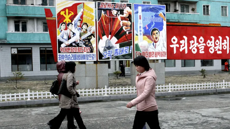 Text na plakátech (zleva): "Vzhůru k úplnému vítězství pod vedením velké strany!", "Nikoliv slovy, ale zbraněmi!" a "Výše, rychleji!" Rudý transparent hlásá: "Posilme a rozšiřme naši stranu jako stranu Kim Ir-sena a Kim Čong-ila!"