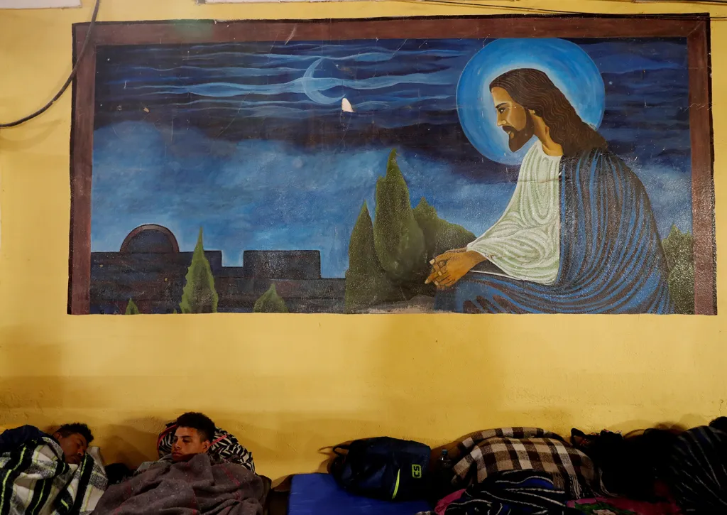 Migranti z tzv. karavany cestující ze Střední Ameriky do Spojených států spí pod náboženskou malbou na zdi v ulicích města Mexicali, Mexiko.