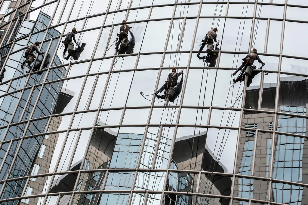 Tvary protějších kancelářských budov se odrážejí ve skleněných tabulích oken, které leští čistící jednotka v Pekingu