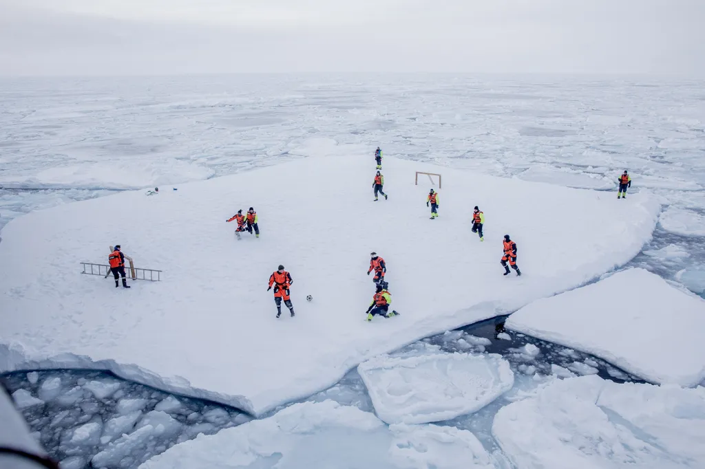 Špicberská posádka norských námořníků a vědců hraje fotbal na velké ledové kře, zatímco ozbrojení strážci zajišťují ochranu před ledními medvědy.