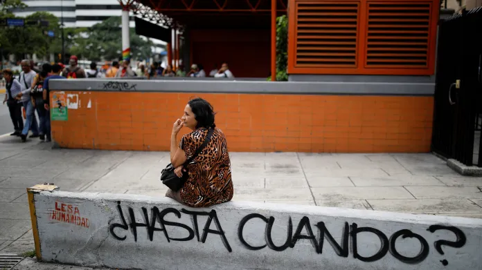 Žena čeká na dopravu vedle graffiti, která v překlaadu zní „Jak dlouho ještě?“ před uzavřenou stanicí metra během výpadku proudu v Caracasu