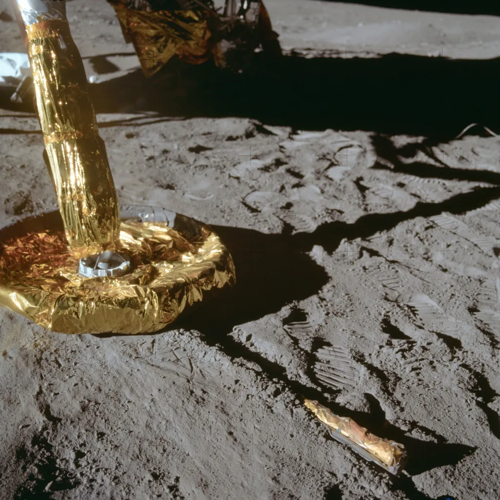 Pohled na podnožku lunárního modulu Apollo 11, spočívající na povrchu Měsíce těsně po přistání. Vyčnívající předmět s tyčinkou je sonda pro snímání povrchu na Měsíci