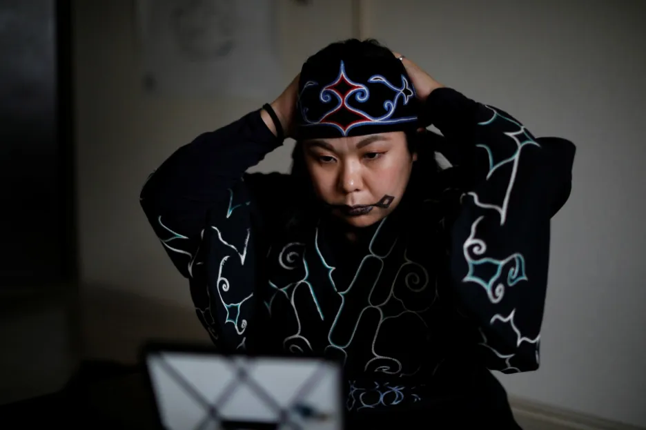 Mai Hachiya nosí tradiční oblečení. Na ústech má rtěnkou namalovaný vzor, který si Ainuové tetují dřívkem namočeným v inkoustu