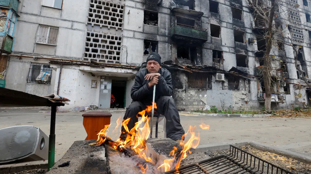 Obyvatel Mariupolu sedí u ohně před těžce poškozeným bytovým domem