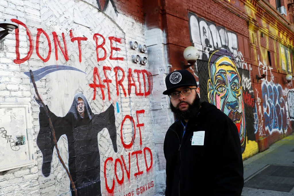 Muž jde kolem stěny, na které je graffiti a nápis sdělující kolemjdoucím, že se nemají covidu obávat. Fotografie je z Houston Street v New Yorku