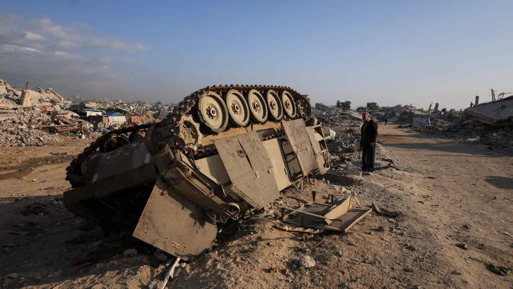 Palestinec stojí vedle zbytků obrněného transportéru a zničených budov v Gaza City (12. 11. 2025)