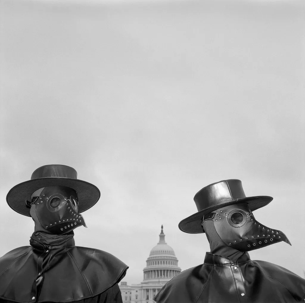 Vítězná fotografie v kategorii Severní a Střední Amerika – dlouhodobý projekt. Pro-očkovací aktivisté nosící masky se zobáky podobné těm, které nosili lékaři ze 17. století během moru. Snaží se upoutat pozornost diváků v okolí Capitol Hill, kde propagovali poselství, že odmítnutí očkování prodlouží pandemii covidu