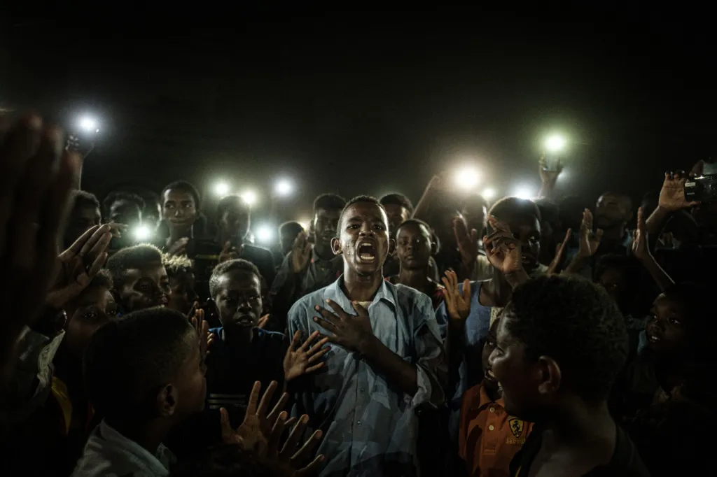 Vítězná fotografie PHOTO OF THE YEAR a GENERAL NEWS: Straight Voice (Přímý hlas). Mladý muž ve světle mobilních telefonů recituje na protest báseň během blackoutu v súdánském Chartúmu. Demonstranti kolem něj skandují hesla volající po civilní vládě
