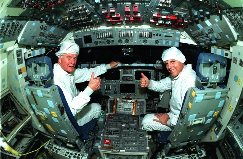 Interiér vesmírného plavidla a v něm sedí senátor John Glenn a astronaut Stephen Oswald v roce 1998