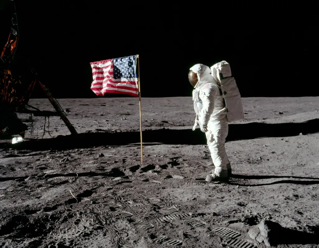 Buzz Aldrin, pilot lunárního modulu, pózuje na fotografii vedle nasazené vlajky Spojených států během extravehikulární aktivity. LM je vlevo a stopy astronautů jsou viditelné v prachu před modulem
