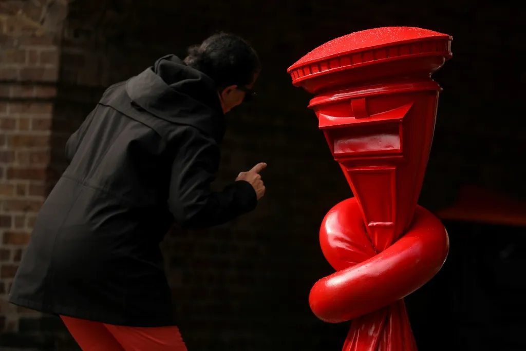 Žena na ulici pozoruje zauzlovanou poštovní schránku. Během festivalu umění v londýnských ulicích tuto sochu představil Alex Chinneck. Dílo nese název „Alphabetti spaghetti“
