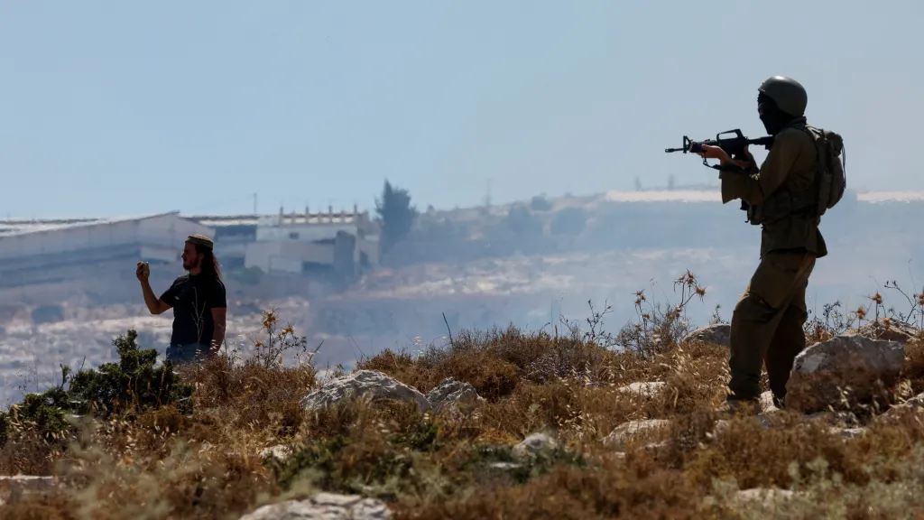 Izraelský osadník (vlevo) natáčí palestinskou demonstraci, zatímco izraelský voják stojí na stráži (okupovaný Západní břeh, 9. června 2026)