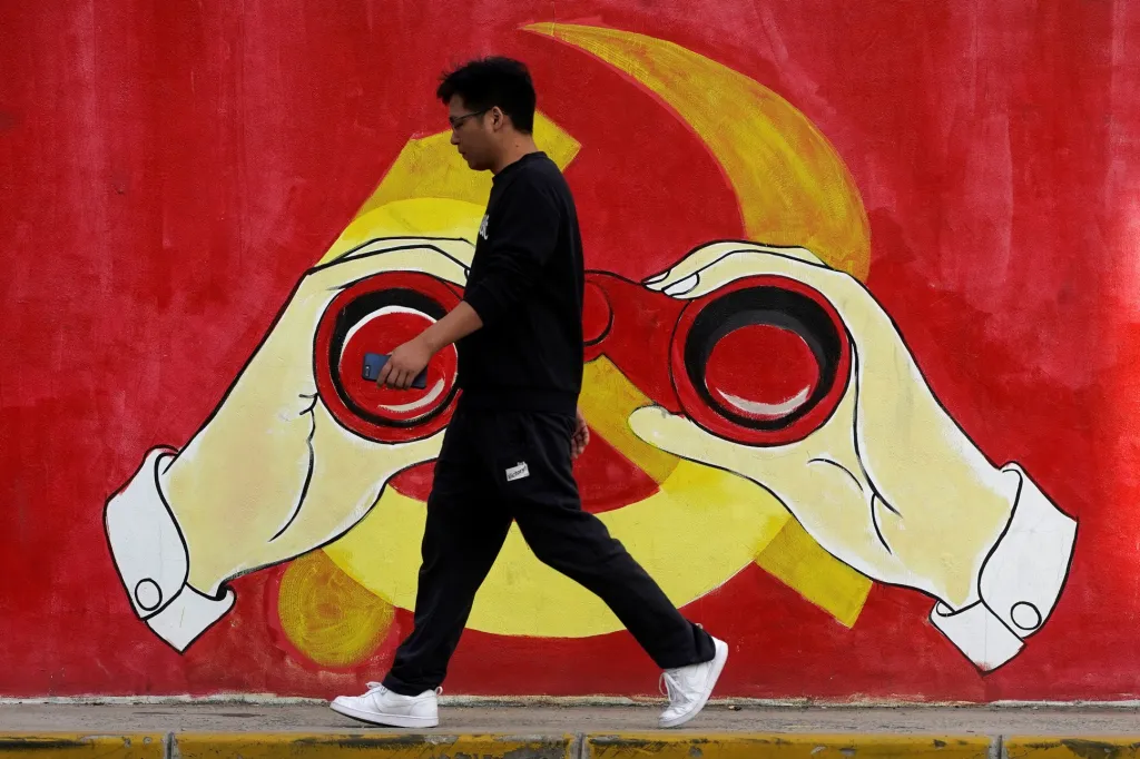 Muž jde podél stěny s graffiti znaku Čínské komunistické strany, který symbolicky vše sleduje. Šanghaj, Čína