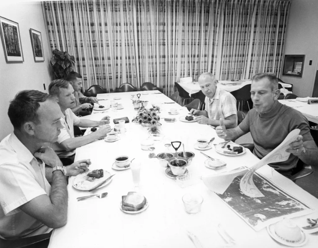 Vedoucí astronaut a ředitel leteckých posádek Donald K. Slayton (vpravo) probírá při snídani měsíční mapy s astronauty Apolla 11 Michaelem Collinsem (vlevo), Neilem Armstrongem a Edwinem Aldrinem (vedle Slaytona). Diskuse se rovněž účastní William Anders (vlevo vzadu), pilot lunárního modulu pro misi Apolla 8