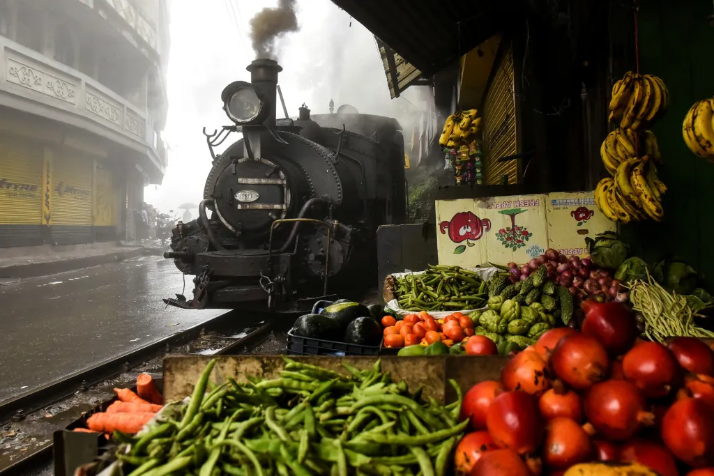 Toto není historie železnic, ale současná fotografie funkční lokomotivy, která stále vozí cestovatele po Darjeeling Himalayan Railway. Na snímku zastavuje před tržištěm ve městě Ghum v Indii.