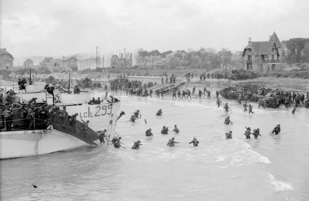 Kanadští vojáci se brodí na břeh po invazi v Bernieres-sur-Mer  6.června 1944. Budova vlevo je nyní známá jako Queen's Own Rifles