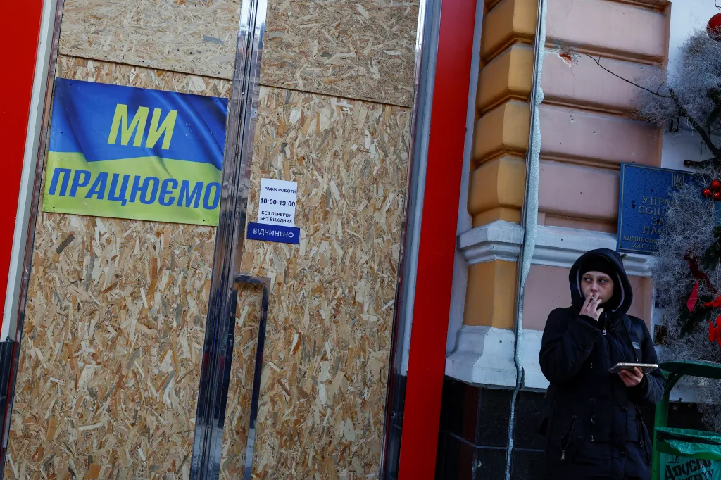 Obyvatelka Charkova kouří vedle obchodu s oblečením, který byl zničen během ruského útoku. Kolemjdoucí si mohou všimnout nápisu hlásící „Máme otevřeno“ (Foceno 13. ledna 2024)