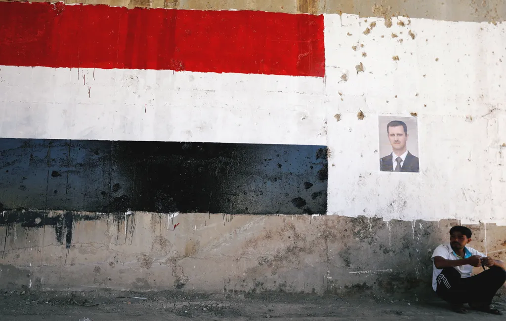 Muž sedící pod nástěnnou malbou syrské vlajky a syrského prezidenta Bašára Asada během znovuotevření silnice mezi městy Homs a Hama