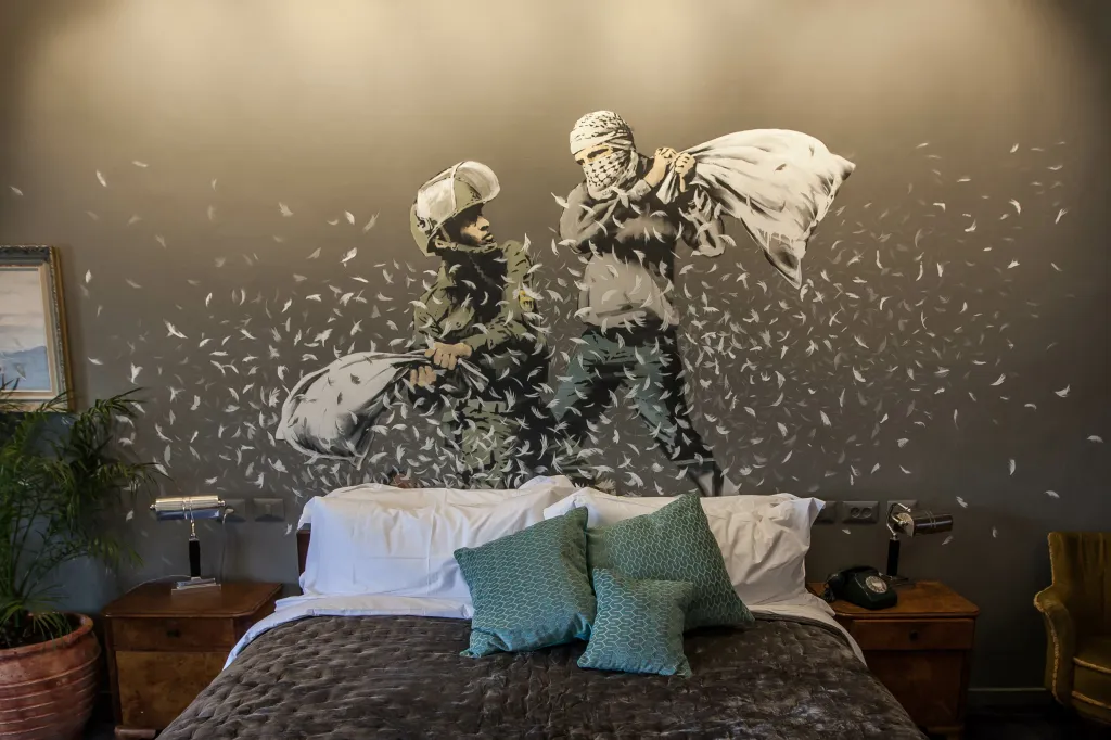 The Walled Off Hotel vyzdobil Banksy svými díly, sžíravý komentář najdou hosté v každém pokoji. Třeba polštářovou bitvu mezi izraelským vojákem a palestinským mladíkem