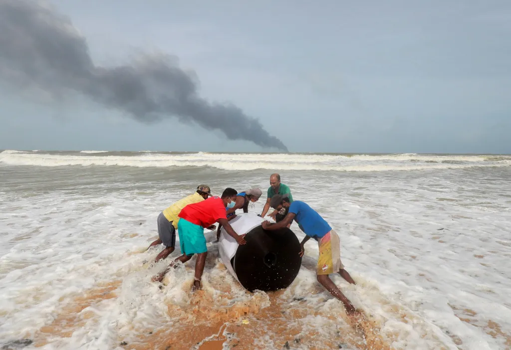 Lidé odklízejí z pláže v Ja-Ela na Srí Lance vyplavený náklad z havarované kontejnerové lodi MV X-Press Pearl. V povzdálí je zachycen kouř, který stoupá z postiženého plavidla