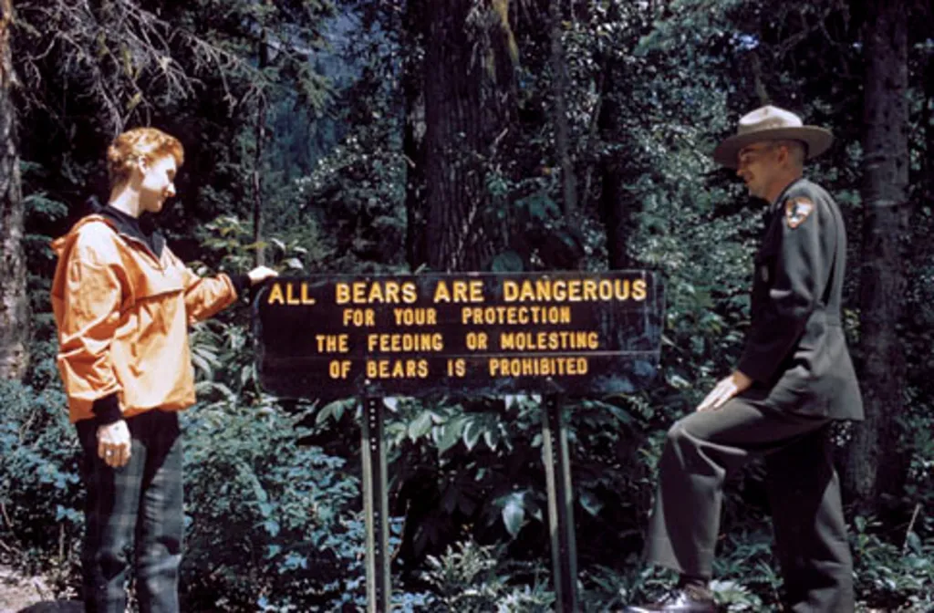 Návštěvnice a strážce parku si prohlíží ceduli s popisem, který zmiňuje nebezpečí před medvědy v Národním parku Glacier v Montaně na fotografii z roku 1958