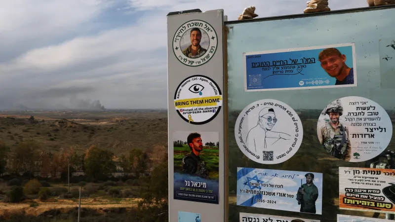 Nálepky padlých izraelských vojáků na zdi v jižním Izraeli, v pozadí stoupající kouř z Pásma Gazy