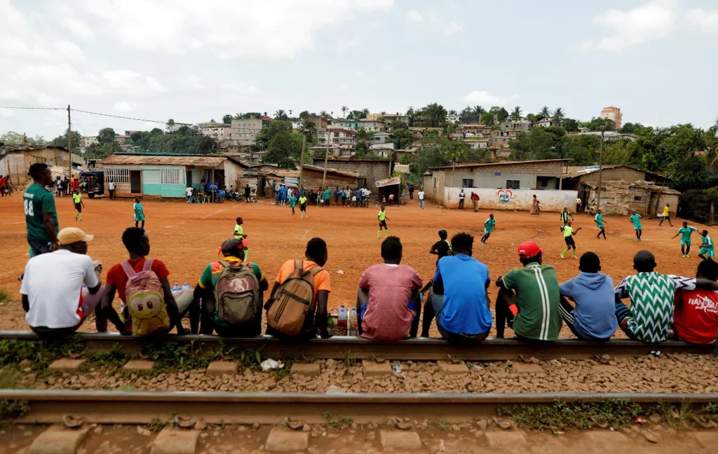 Převážně mužské publikum sleduje přátelský zápas mezi týmem chlapců U15 a týmem dívek U17, který je trénovaný profesionálními trenéry na hřišti RFA v Yaoundé