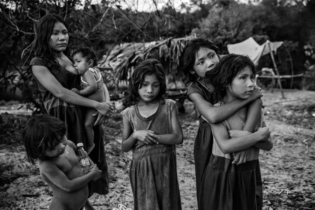 Vítězná fotografie v kategorii Dlouhodobý projekt. Ženy a děti z komunity Pirahã stojící vedle svého tábora na břehu brazilské řeky Maici v roce 2016. Sledují řidiče projíždějící po transamazonské dálnici a doufají, že dostanou nějaké jídlo nebo nápoje