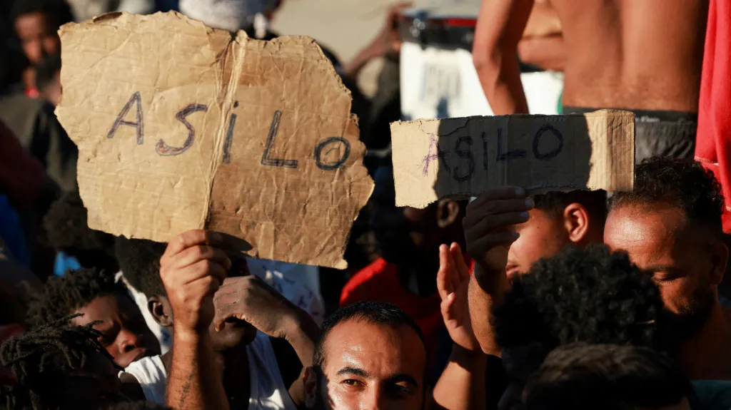 Migranti drží nápis Azyl, zatímco čekají v Ceutě na jídlo a pití od Červeného kříže (3. srpna 2026)