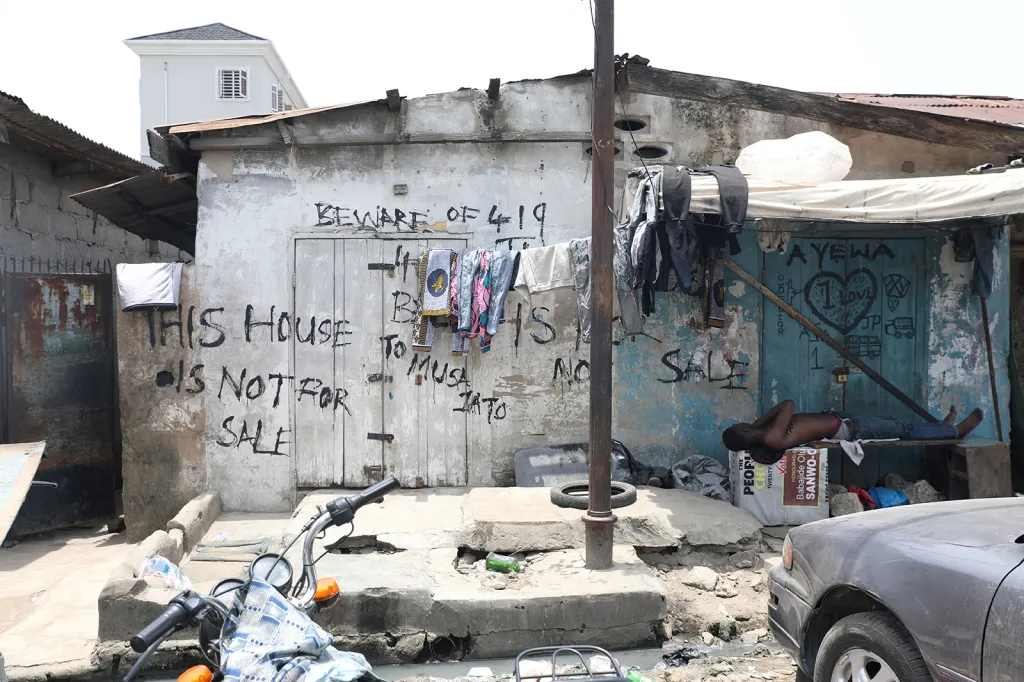 Muž při odpoledním odpočinku na lavičce před zchátralou budovou s nápisem „Tento dům není na prodej“ ve městě Lagos v Nigérii