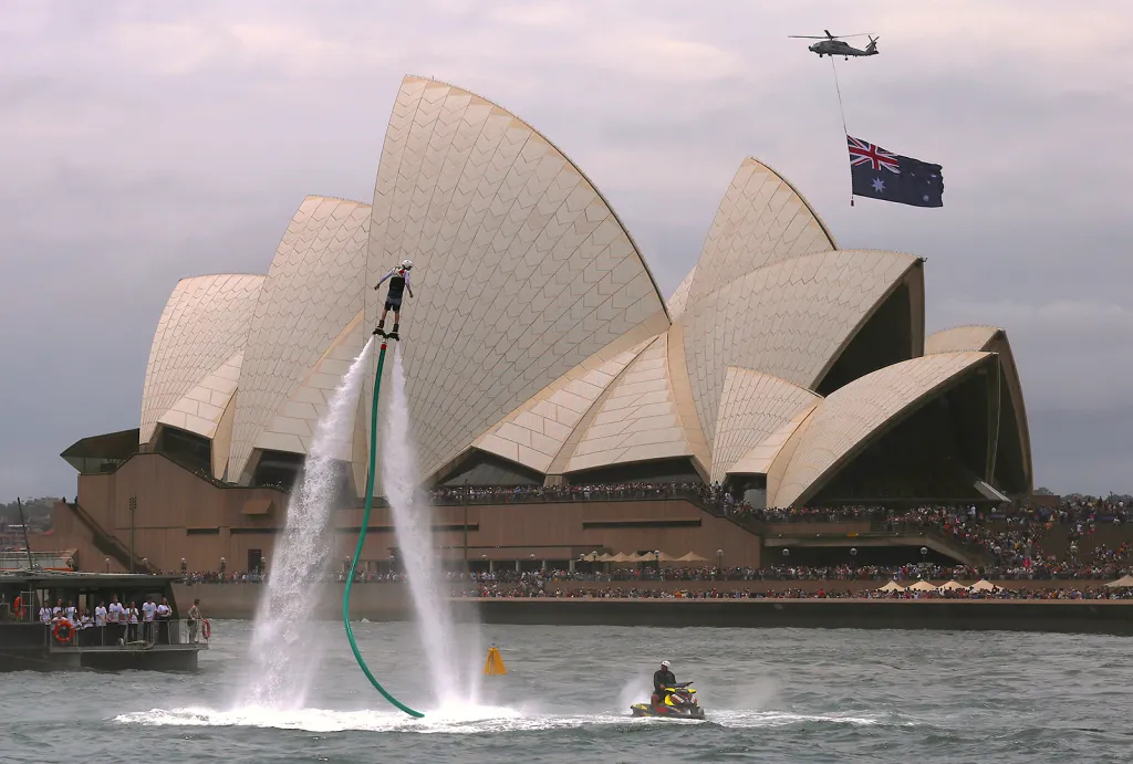 Australská vlajka pod vojenskou helikoptérou nad Operou v Sydney a performance flyboardu během oslav Dne Austrálie, který připomíná první příjezd Britů v roce 1788