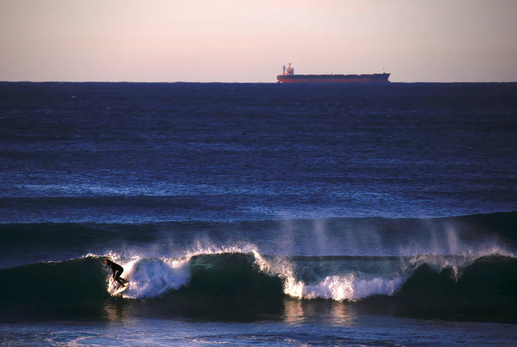Surfař na vlně u pláže Merewether v australském Newcastlu a nákladní loď na obzoru čekající na svůj náklad uhlí.